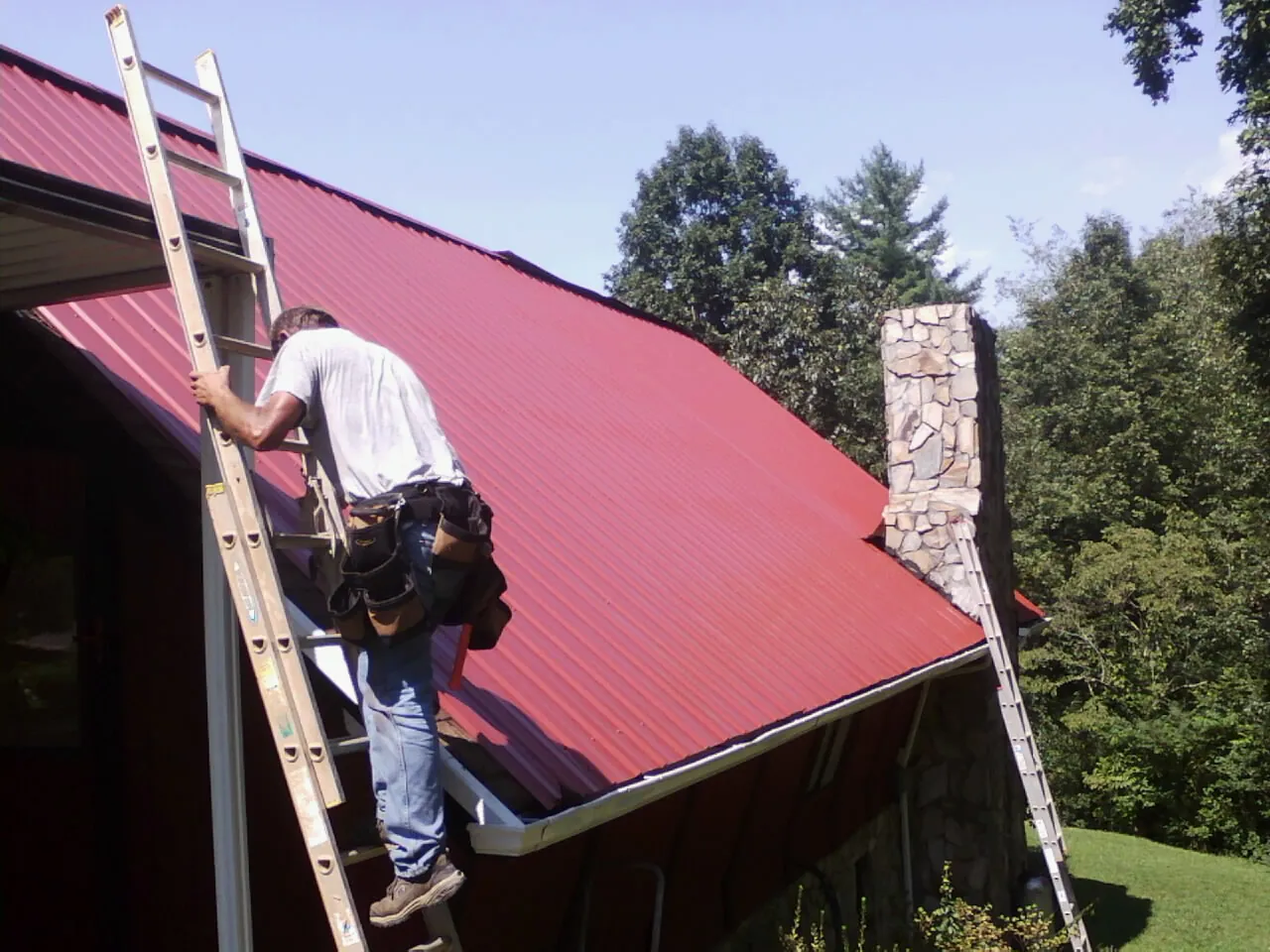 Roofer on a ladder beside a red metal roof and stone chimney — Soffit & Fascia Repair by Roofing Brothers in Allamuchy